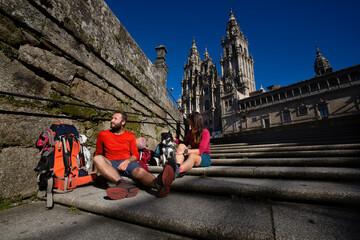Two pilgrims finish the Camino de Santiago and rest on the steps of Plaza del Obradoiro in front of the Cathedral of Santiago de Compostela - obrazy, fototapety, plakaty