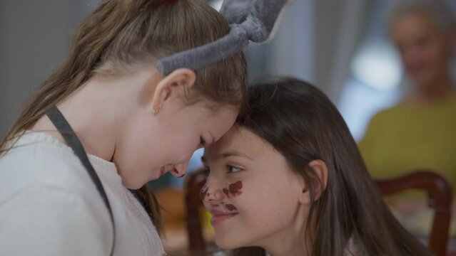 Close-up joyful teenage and little sisters rubbing noses smiling with colored faces indoors. Happy cheerful siblings having fun at home on Easter Sunday with blurred grandmother at background