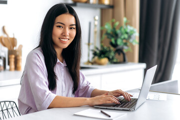 Positive pretty successful Chinese brunette girl, designer, freelancer or student, in shirt, sits at home in the kitchen with a laptop, remote work or study, looking at the camera, smiling friendly