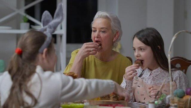 Happy joyful grandmother laughing talking with granddaughters eating Easter dinner at table indoors. Portrait of gorgeous Caucasian woman enjoying leisure with girls on holiday at home