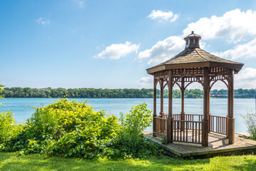 A wood gazebo over looks the Niagara River in Niagara on the Lake, Ontario, Canada on a blue sky day. The United States can be seen in the distance. - obrazy, fototapety, plakaty