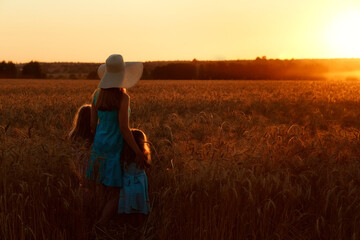 A mother and daughter in a wheat field look at the sun. A woman in a hat and a dress with a girl enjoying the sunset in nature.