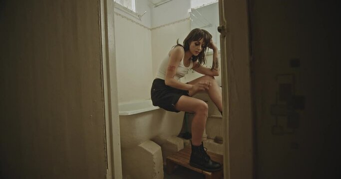 Dejected woman sitting smoking on an old bathtub indoors. Dejected woman with tattoos sitting smoking on an old bathtub indoors viewed through the door