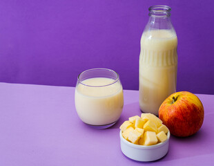 Glass bottle and large glass with milk and a whole red apple next to a bowl with apple cubes in a purple or violet environment. Copy space.
