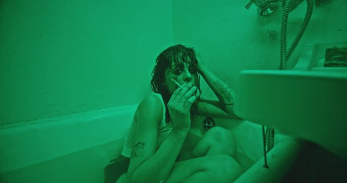 Depressed female junkie smoking in bathtub. Zoom out view of unhappy addicted woman with smeared makeup touching wet hair and smoking marijuana joint while sitting in bathtub with water in bathroom