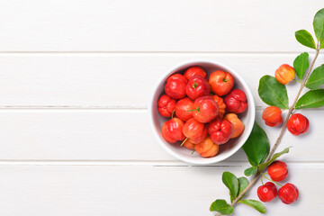 Naklejka na ściany i meble Flat lay of Acerola cherry in white bowl with branch on white wood.