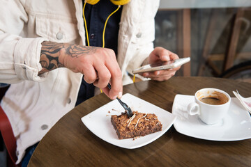 Close-up of an unrecognizable man eating a cake and drinking coffee while checking his smart phone