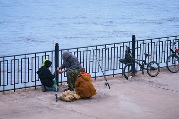 An adult fisherman gives bait for fishing to two boys on the city embankment. Fishing rods and...