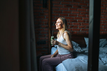 Fototapeta na wymiar Young adult woman with blonde long hair in casual with green smoothie near window at home