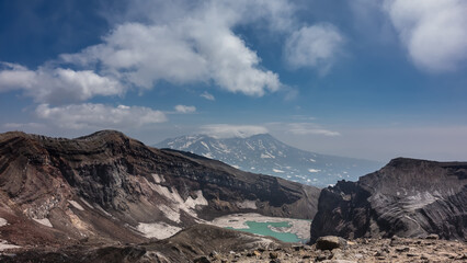 Turquoise acid lake on top of an active volcano. Melted snow at the water's edge. The layered structure of steep mountain slopes is visible. Clouds in the blue sky. Kamchatka. Gorely Volcano