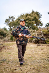 Full-body male hunter in camouflage outerwear and cap carrying a gun ready to shoot