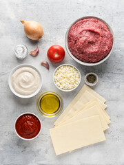 Lasagna ingredients top view. Ground beef (minced), lasagne sheets, mozzarella cheese, tomato, bechamel sauce, olive oil, onion bulb, garlic cloves, basil. Traditional italian dish. Grey background.
