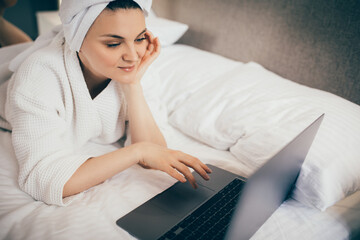 Young woman freelancer working from home using her laptop, lying in bed in white bathrobe and towel on head.