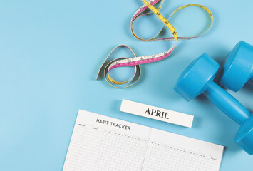  flat lay of habit tracker book, wooden calendar April, blue  dumbbells and measuring tape on blue...