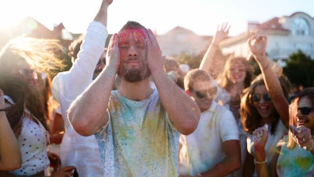 Joyful man rubs colored powder in face and beard, jumps into friends hands imitating stage diving. Friends have fun at hindu holiday of spring, colors. End of covid pandemic isolation. Slow motion.