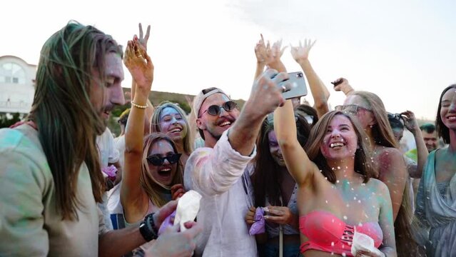 Cheerful people throw colorful powder, take selfie on smartphone and smile at Holi festival on beach in slow motion. Friends smeared in dry colors take group photo on phone. End of covid pandemic