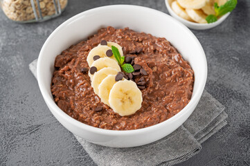 Chocolate oatmeal porridge with banana and chocolate chips on top in a white bowl. Healthy breakfast. Copy space.