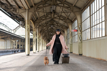 Young cute girl with dog and suitcases on platform waiting for train in pink coat, at the Vitebsk...