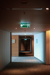 Emergency exit sign at the corridor in building. Green fire exit sign hanging on ceiling on dark corridor in building near fire emergency exit door. Green emergency exit sign.