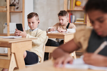 Wide angle portrait of teenage boy using smartphone in school while sitting at desk in classroom, copy space