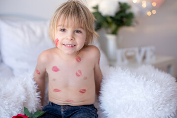 Beautiful blond toddler child, boy with lipstick kisses on his body, holding red rose for Valentine