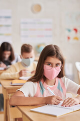 Vertical portrait of girl wearing mask in school and looking at camera while sitting at desk in classroom
