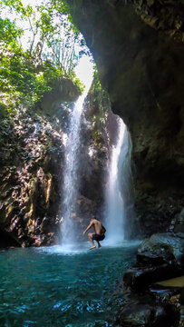 A man in swimsuit jumping from the slippery rocks in front of Gitgit Twin Waterfall, Bali, Indonesia. The waterfall is surrounded by rocks. The man enjoying the beauty of the nature. Adventure
