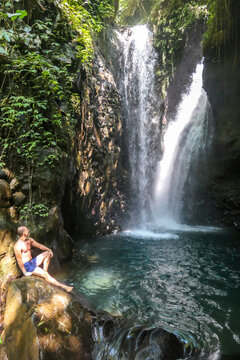 A man in swimsuit sitting on the rocks in front of Gitgit Twin Waterfall, Bali, Indonesia. The waterfall is surrounded by rocks. The man enjoying the beauty of the nature. Adventure and discovery