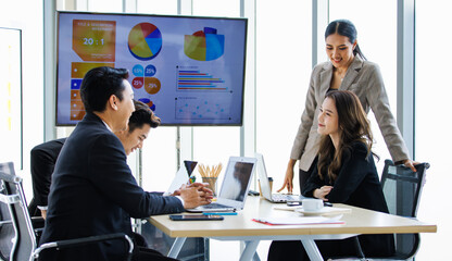 Asian professional successful businesswoman in formal suit standing showing presenting explaining report investment graph chart data from computer monitor in meeting room to business team colleagues