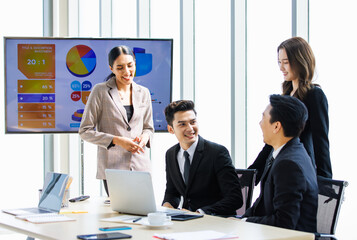 Asian professional successful businesswoman in formal suit standing showing presenting explaining report investment graph chart data from computer monitor in meeting room to business team colleagues