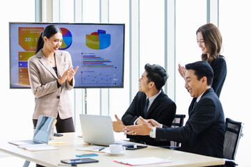 Asian professional successful businesswoman in formal suit standing showing presenting explaining report investment graph chart data from computer monitor in meeting room to business team colleagues