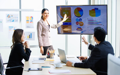 Asian professional successful businesswoman in formal suit standing showing presenting explaining report investment graph chart data from computer monitor in meeting room to business team colleagues