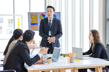 Asian professional successful businessman in formal suit standing showing presenting explaining report investment graph chart data from computer monitor in meeting room to businesswoman colleagues