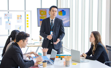 Asian professional successful businessman in formal suit standing showing presenting explaining report investment graph chart data from computer monitor in meeting room to businesswoman colleagues