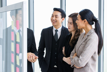 Asian happy male and female professional successful businessman and businesswoman colleagues partnership teamwork in formal business suit standing smiling look at post it note on glass board together