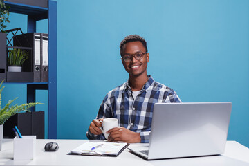 Joyful smiling heartily office worker having cup of coffee while sitting in professional interior. Cheerful businessman in research company office sitting at desk while looking at camera.