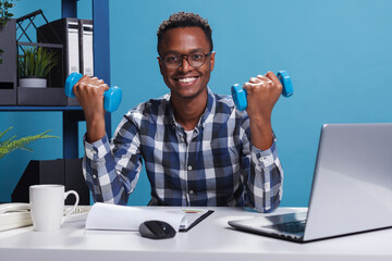 Company athletic businessman lifting weights while sitting at desk in office. Active and healthy young adult doing workout with fitness dumbbells in order to maintain body strong.