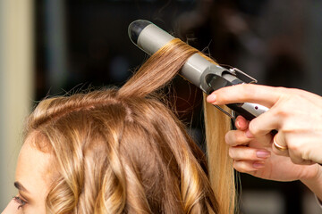 Fototapeta premium Hairdresser makes curls with a curling iron for the young woman with long brown hair in a beauty salon