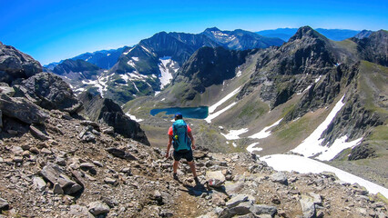 Fototapeta premium A young woman with a big backpack hikes down towards a clear, navy blue lake hiding between tall mountain peaks. Some of the slopes are covered with snow. In the back is another mountain range