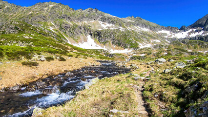 Fototapeta na wymiar A serene view on snowy mountain from a small stream's side. The stream starts it's long way to the sea. Tall glacier towering above the landscape. Spring in the alpine valley.