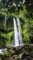 Obraz premium A man in swimsuit standing under two levelled waterfall in Lombok, Indonesia. Tiu Kelep Waterfall is surrounded by lush green plants from each side. Long and powerful waterfall. Beauty of the nature.