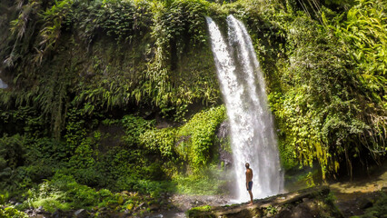A man in swimsuit standing under two levelled waterfall in Lombok, Indonesia. Tiu Kelep Waterfall is surrounded by lush green plants from each side. Long and powerful waterfall. Beauty of the nature.