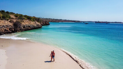 A drone shot of a couple playing at the seashore of Pink Beach in Lombok. Hidden gem, not spoiled by tourists. Waves gently spreading on the beach. Turquoise color of the water. Joyful playtime.