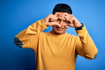 Naklejka premium Young handsome latin man wearing yellow casual sweater over isolated blue background Doing heart shape with hand and fingers smiling looking through sign