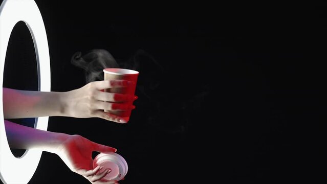 Coffee aroma. Takeout drink. Delivery service. Female hand opening paper cup with hot beverage steam in LED light ring isolated on dark night black empty space advertising background.