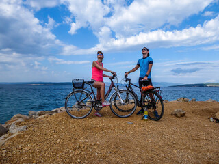 A couple in sporty outfits, standing on their bikes and checking the weather. Behind them there is Mediterranean Sea. Thick clouds above them. Active holidays. Beautiful coastal line of Croatia - obrazy, fototapety, plakaty
