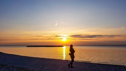 A young woman standing on the beach and watching sunset. The beach is pebbly and overgrown with grass. Sun reflects itself in the calm water of the lake. Sunrise above the lake. Freedom and happiness