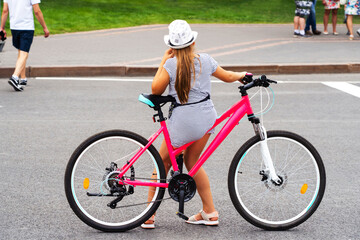 Fototapeta premium A teenage girl in a white hat is resting next to a pink bicycle. Walk on a two-wheeled transport through the city center. Unrecognizable person. Back view