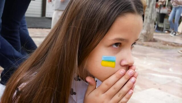 Child with blue and yellow ukrainian flag painted on cheeks sitting on the street.