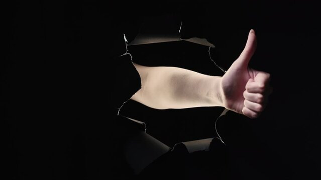 Vote gesture. Yes no. Positive negative feedback. Female hand showing thumb up down in black breakthrough paper hole ripped wall isolated on dark night copy space background loop.